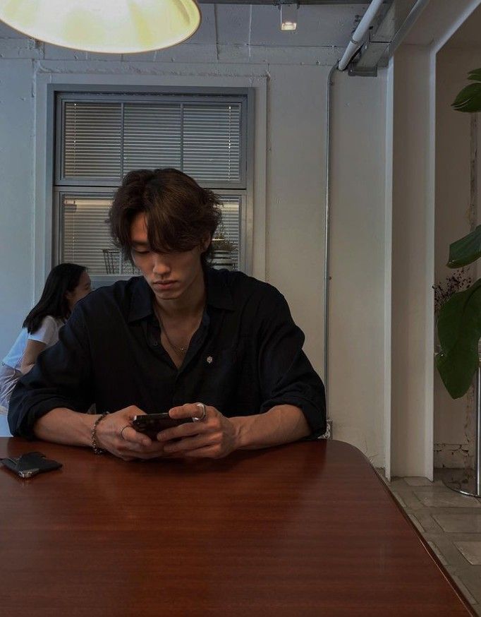 a man sitting at a table looking at his cell phone while another person stands in the background