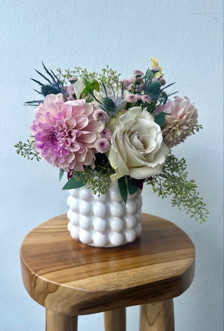 a white vase filled with lots of flowers on top of a wooden stool next to a wall