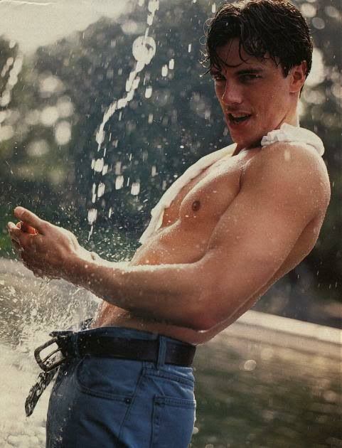 a shirtless man standing in front of a fountain holding his cell phone and smiling