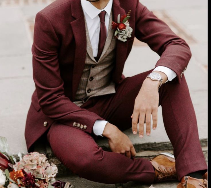 a man in a red suit and tie sitting on the ground next to some flowers