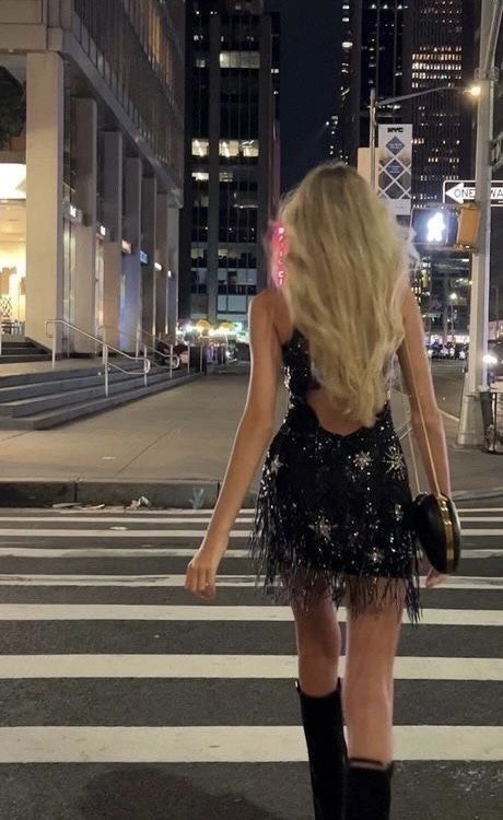 a woman walking across a cross walk in front of tall buildings at night with her back to the camera