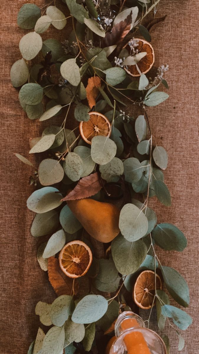 an arrangement of oranges and greenery on a brown cloth with glassware in the foreground