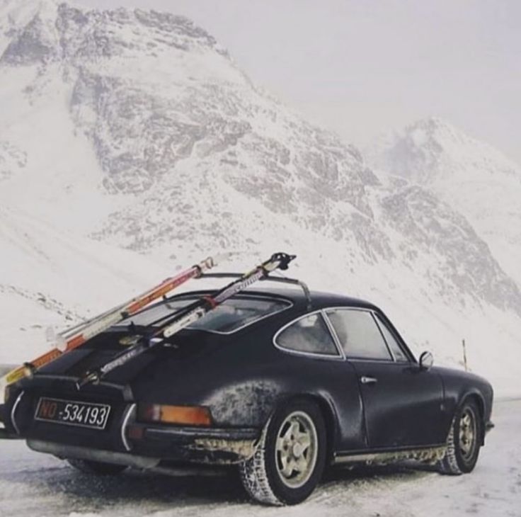 an old car with skis on the roof parked in front of a snowy mountain