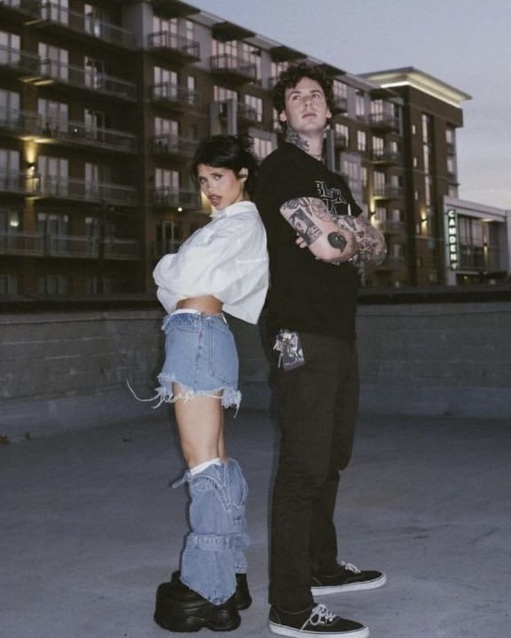 two people standing next to each other with tattoos on their arms and legs, in front of an apartment building