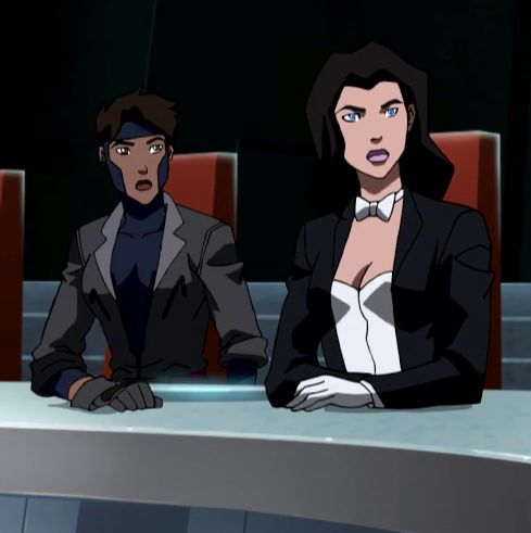 two people sitting at a table in front of red chairs and one person wearing a black suit