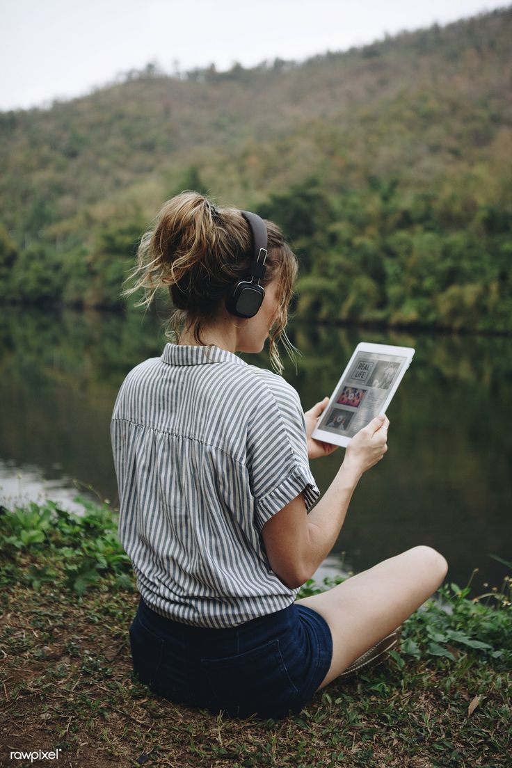 a woman sitting on the ground reading a magazine next to a lake with trees in the background