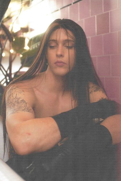 a shirtless man with long hair sitting in a bath room next to a pink tiled wall