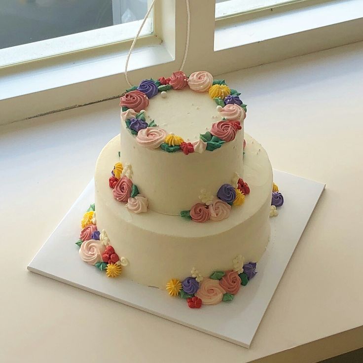 a three tiered white cake with colorful flowers on the top and bottom, sitting in front of a window