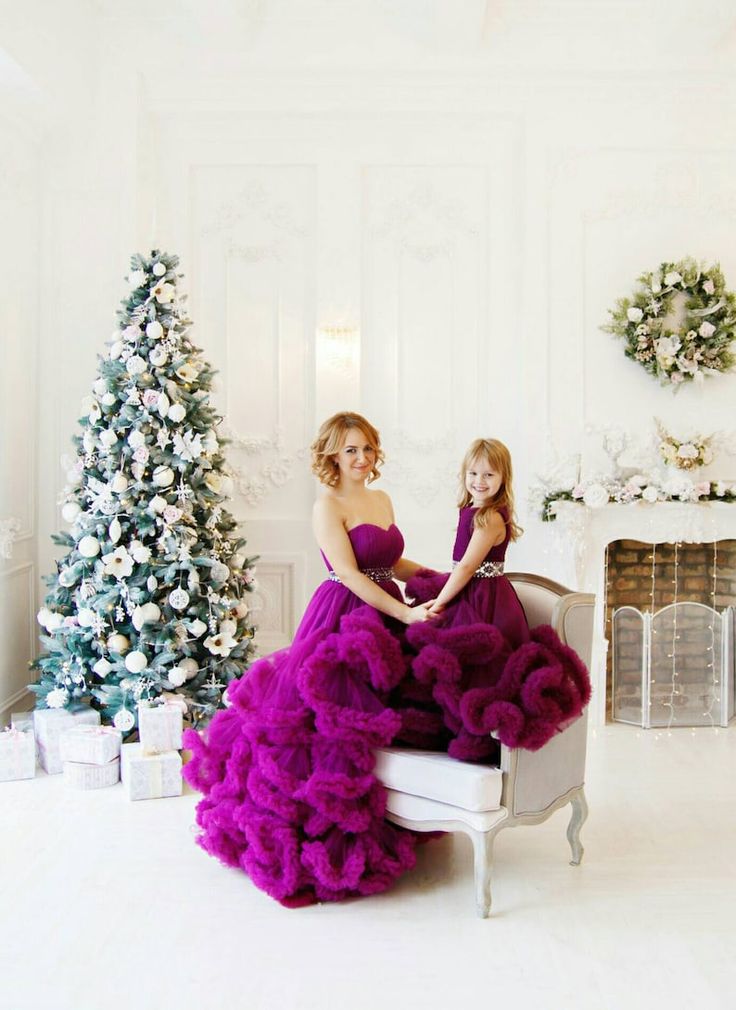 two women in dresses are sitting on a chair with christmas trees behind them and one woman is wearing a purple dress