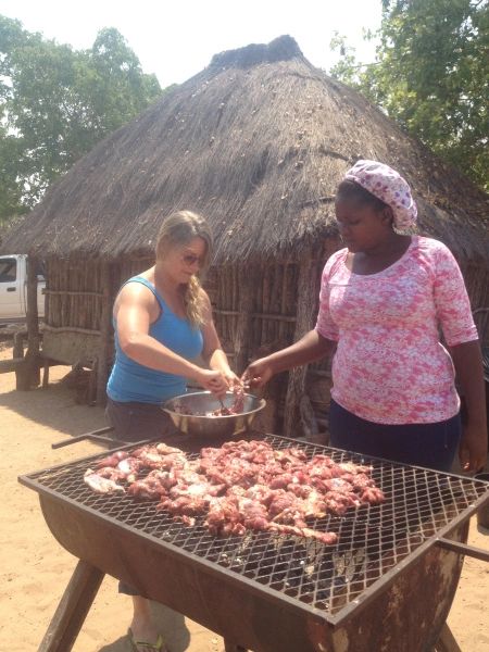 two women grilling meat on an outdoor bbq with a straw hut in the background