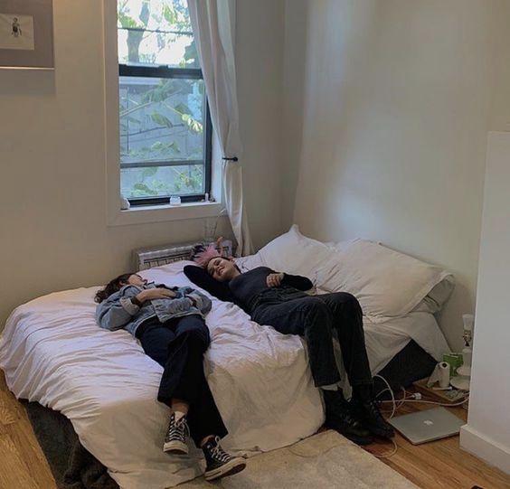 two people laying on a bed in a room with wood flooring and white walls