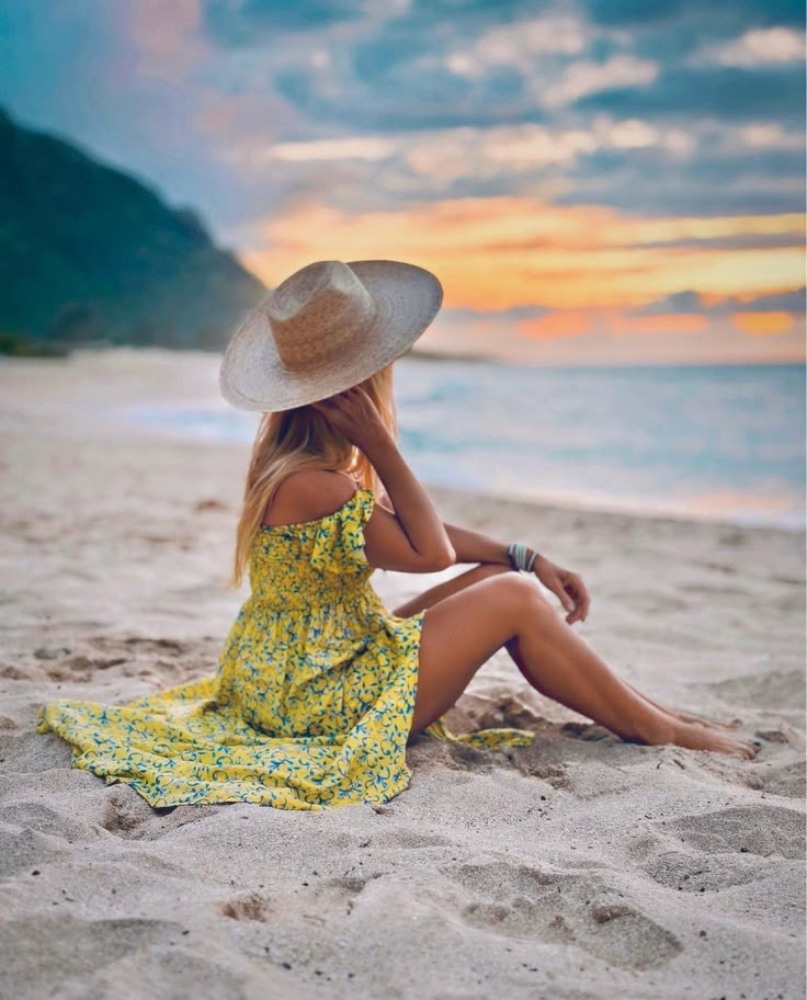 a woman sitting on top of a sandy beach next to the ocean wearing a hat