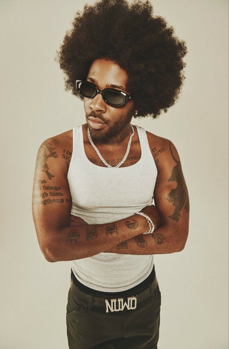 a man wearing sunglasses and a tank top with tattoos on his arms, standing in front of a white background