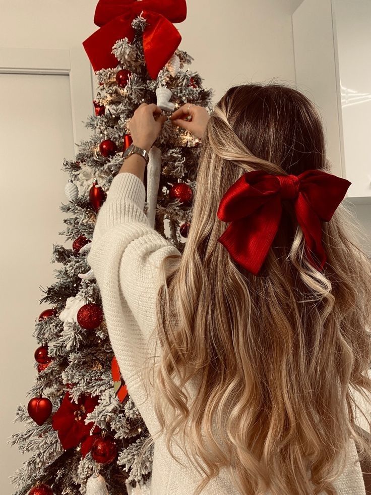 a woman with long blonde hair standing next to a christmas tree and holding a red bow