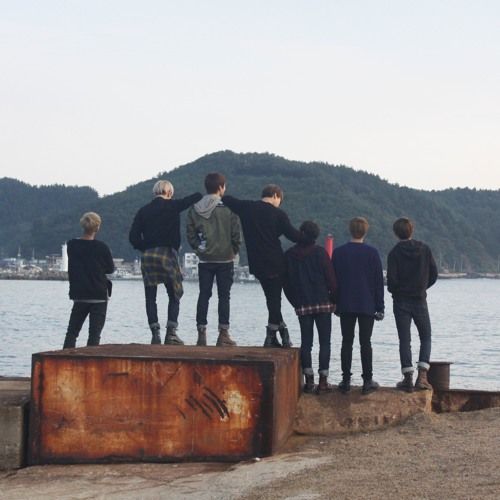 five people standing on top of a piece of wood near the water with mountains in the background