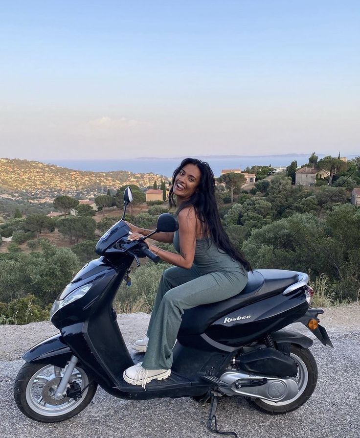 a woman sitting on top of a scooter in front of some trees and hills