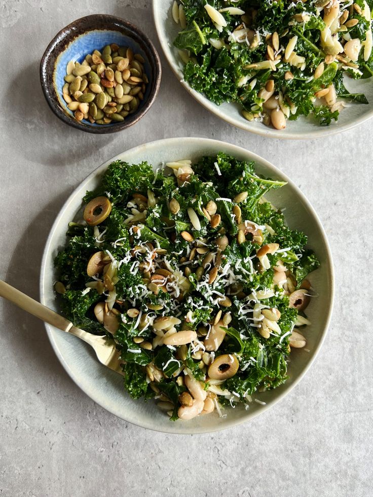 two white bowls filled with greens and nuts on top of a marble countertop next to a wooden spoon