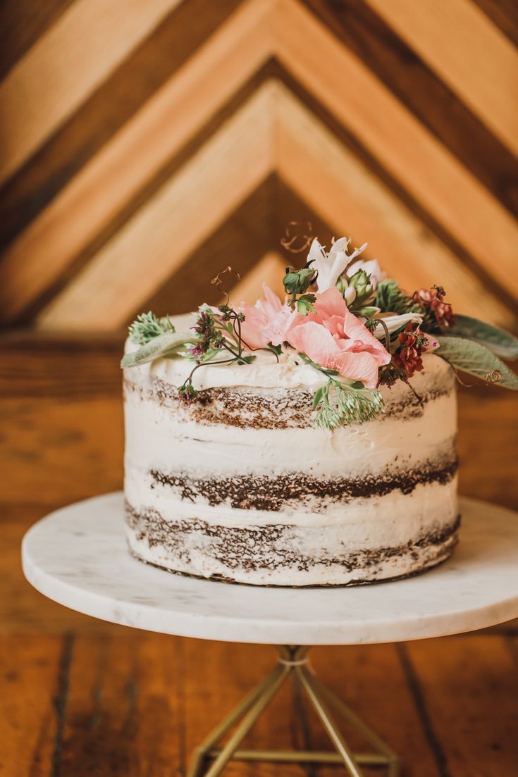 a white cake with flowers on top is sitting on a small metal stand in front of a wooden wall