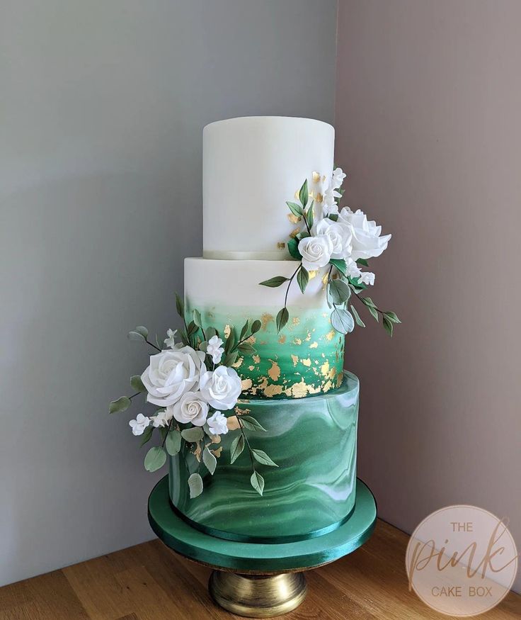 a three tiered cake with white flowers and greenery on the top is sitting on a green stand