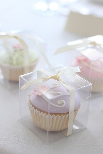 three cupcakes with bows and ribbons in small clear boxes on a white table