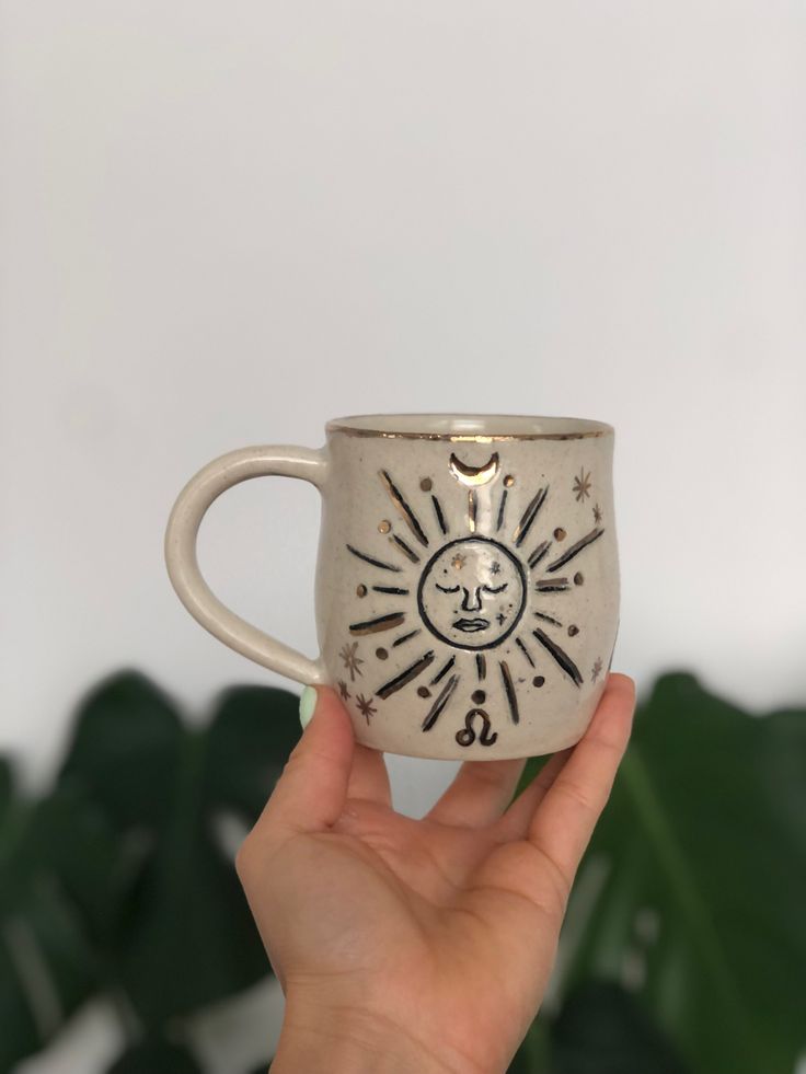 a hand holding a white coffee cup with sun and moon designs on the outside, in front of a green plant