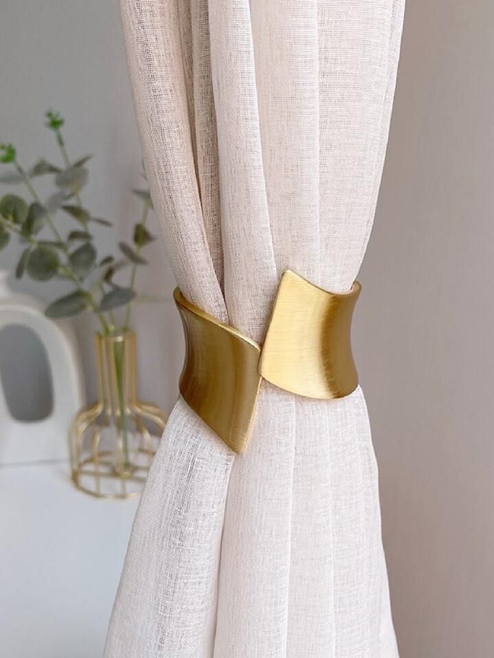 a white curtain with gold colored metal hardware on it's side, next to a potted plant