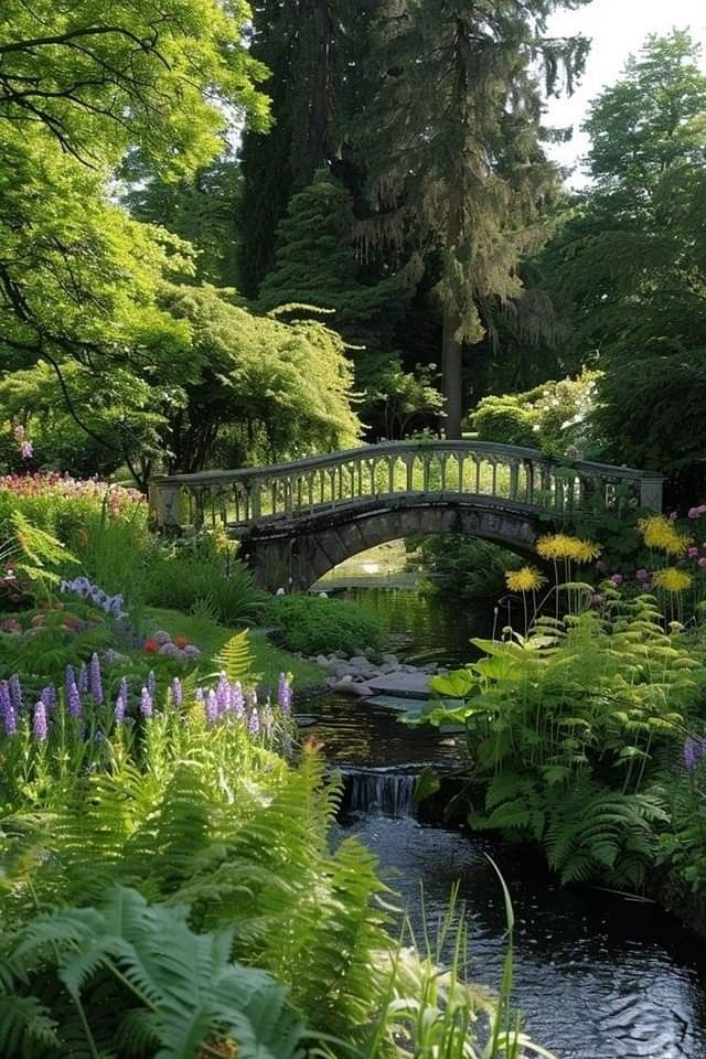 a bridge over a small stream surrounded by lush green trees and flowers in the foreground