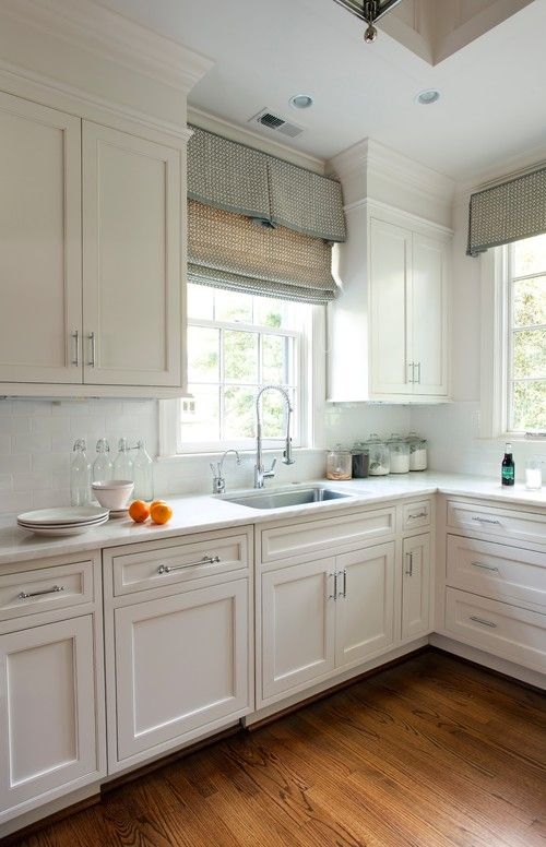 a kitchen filled with white cabinets and wooden flooring next to two window sills