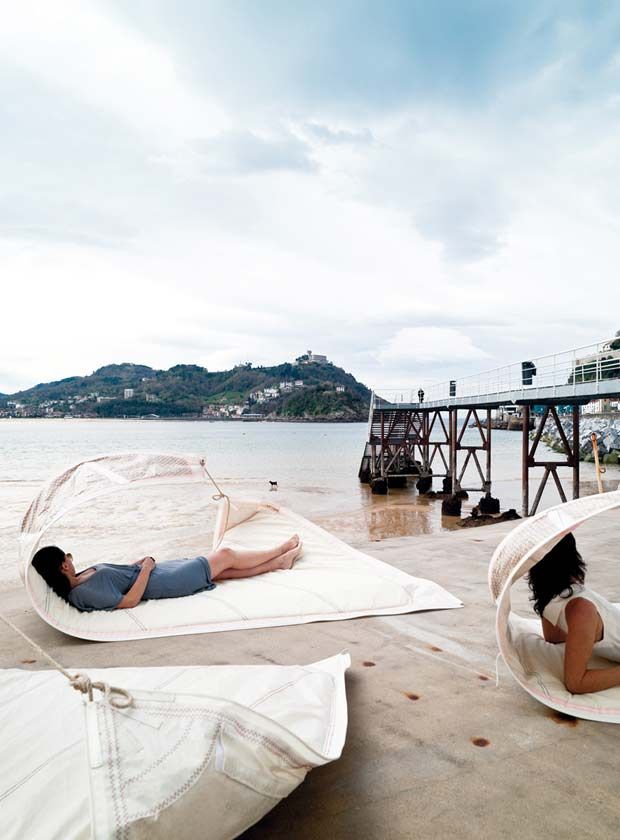 two people laying on mattresses in the sand near water and a pier with boats