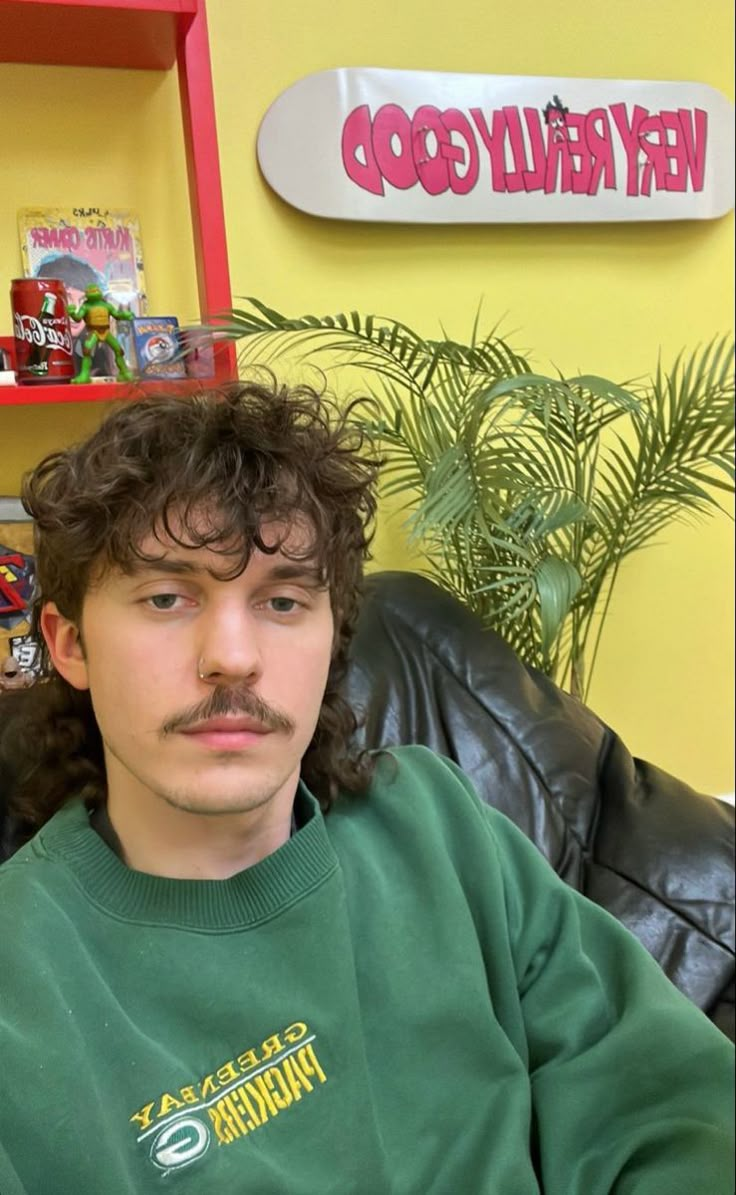 a man with curly hair and a mustache sitting in a black leather chair next to a potted plant