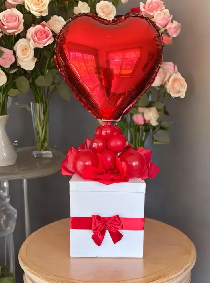 a heart - shaped box with red ribbon and bow sits on a table next to flowers