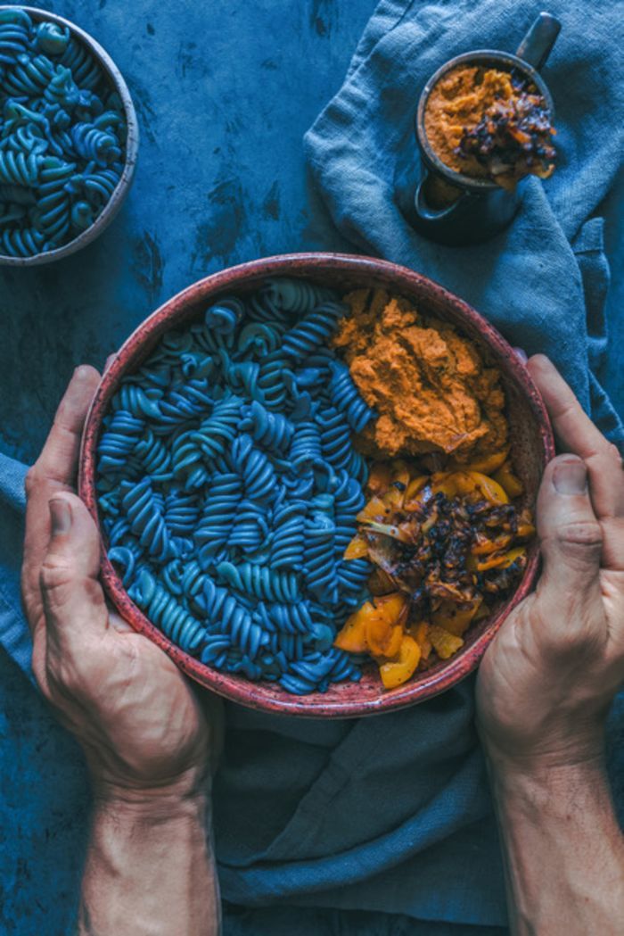 two hands holding a bowl filled with pasta and other food on top of a table