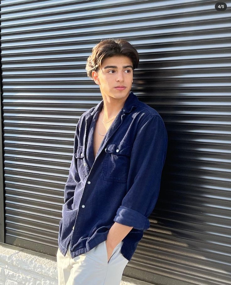 a young man leaning against a wall wearing white pants and a blue button up shirt