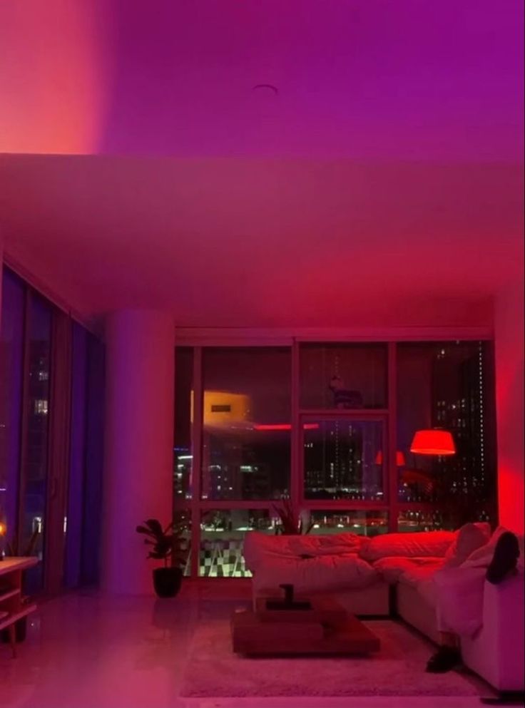 a living room filled with furniture and purple lighting in the dark night time, overlooking a cityscape