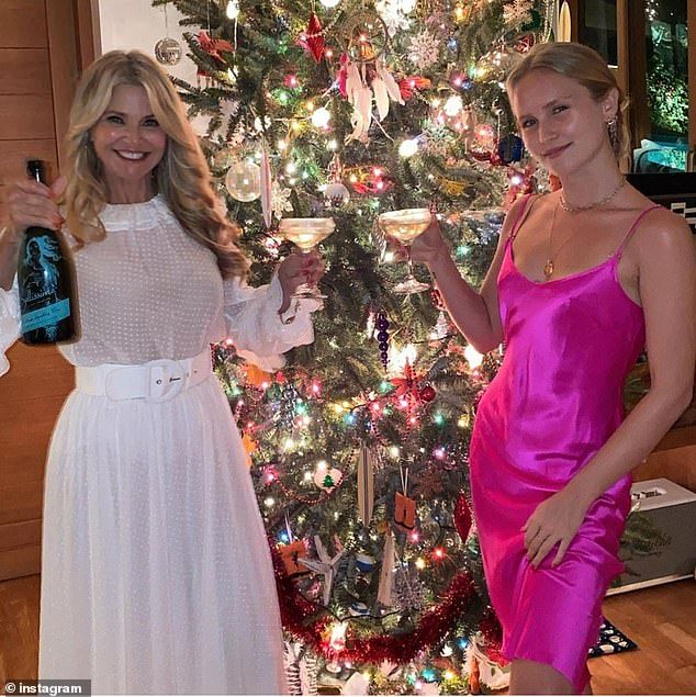 two women standing next to each other in front of a christmas tree holding wine glasses