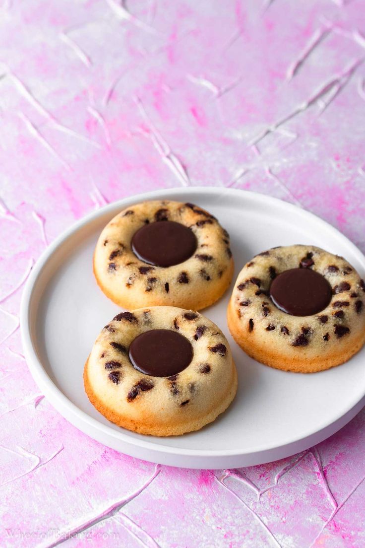 three chocolate chip donuts on a white plate with pink paint splattered in the background