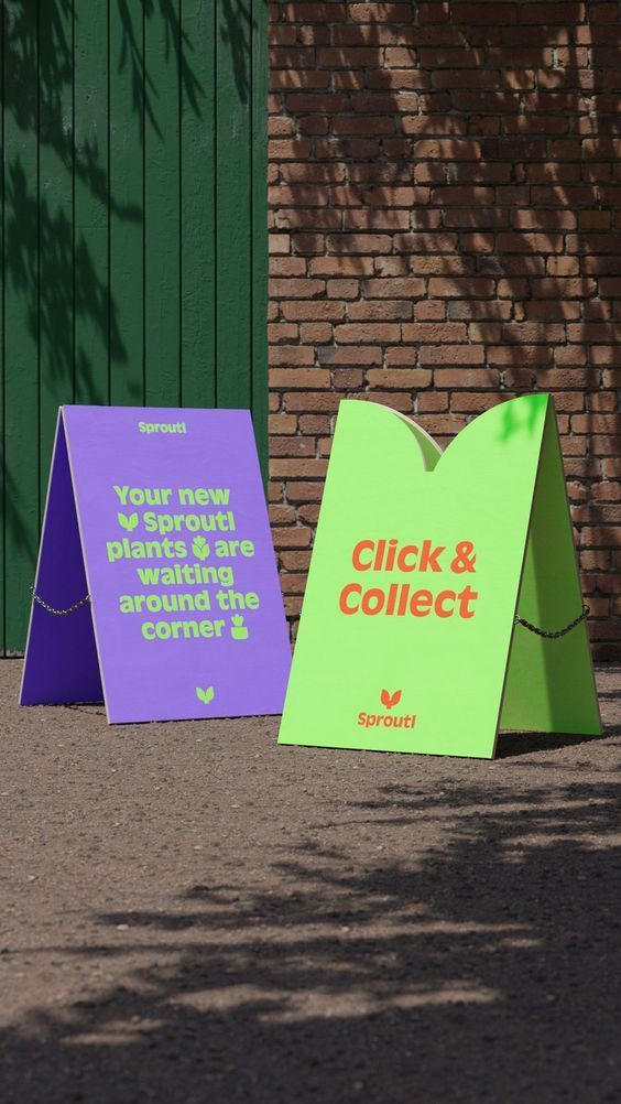 two colorful signs sitting on the side of a road next to a brick wall and tree