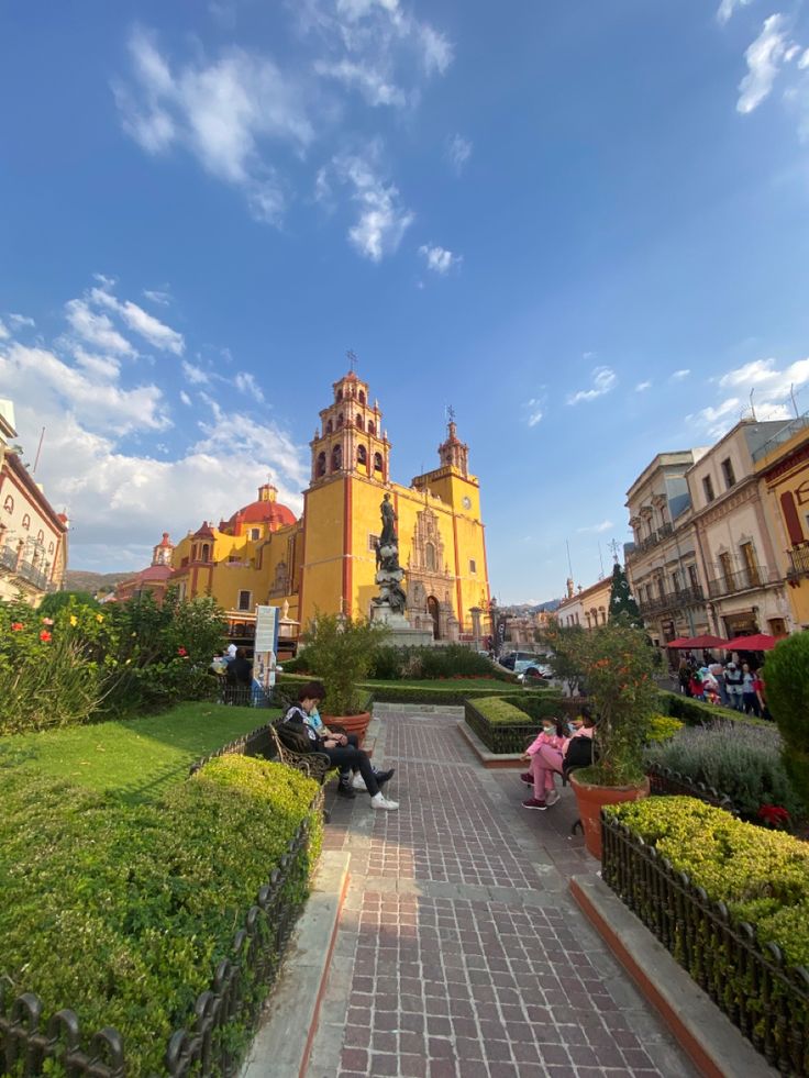 people are sitting on benches in the middle of a walkway near some buildings and bushes