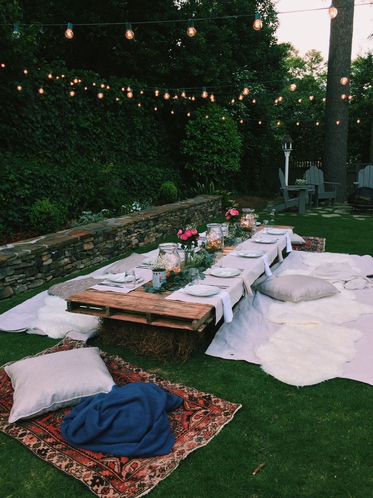 a picnic table set up with pillows, blankets and candles on the grass in front of a stone wall