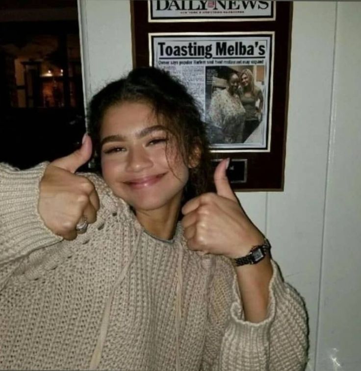 a woman giving the thumbs up sign in front of a news paper advertisement on a wall