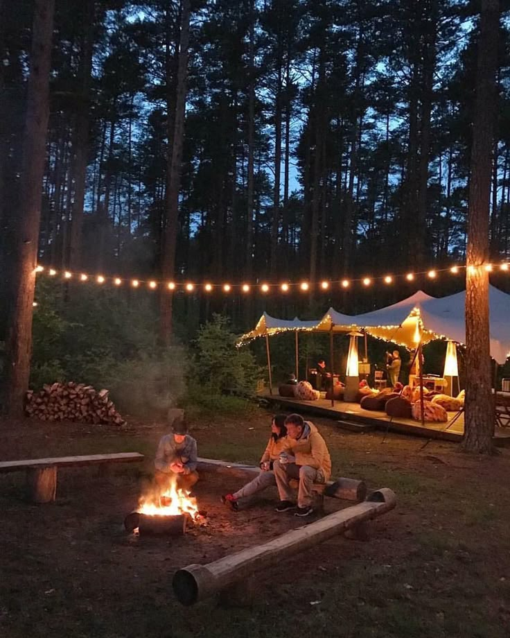 people sitting around a campfire in the woods at night with lights strung over them