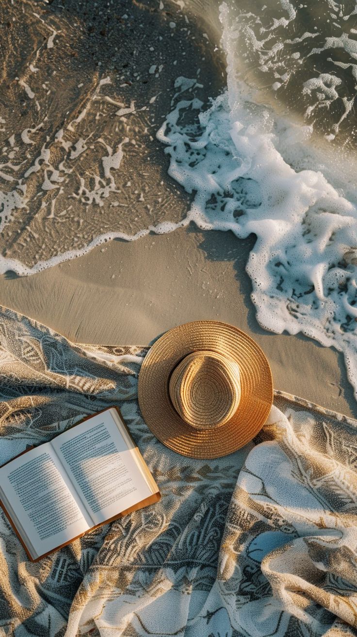 an open book and hat on a blanket near the ocean with waves coming in from the shore