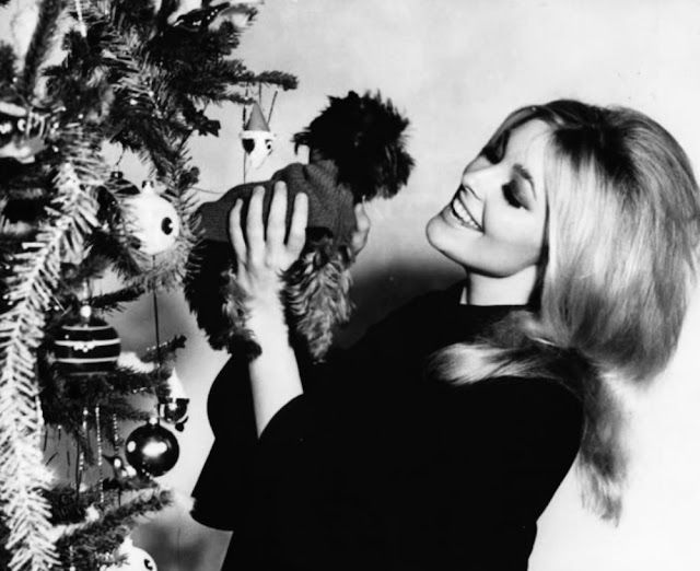 a woman holding a teddy bear next to a christmas tree with ornaments on it's branches