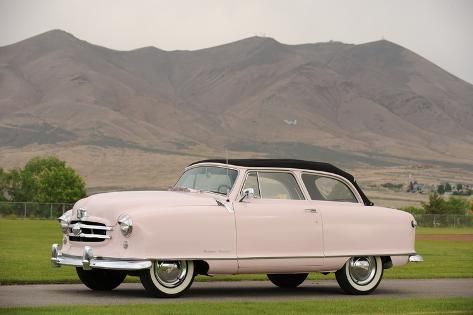 an old pink car is parked on the side of the road with mountains in the background