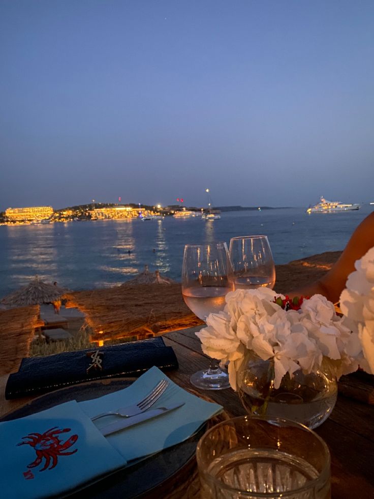 two wine glasses are sitting on a table with flowers in front of the water at night