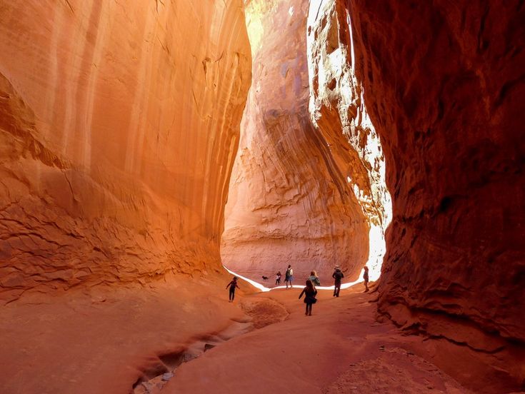 three people are walking through a narrow slot in the rock formation, with sunlight streaming through