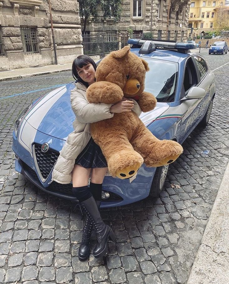 a woman sitting on the hood of a car holding a large teddy bear in front of her