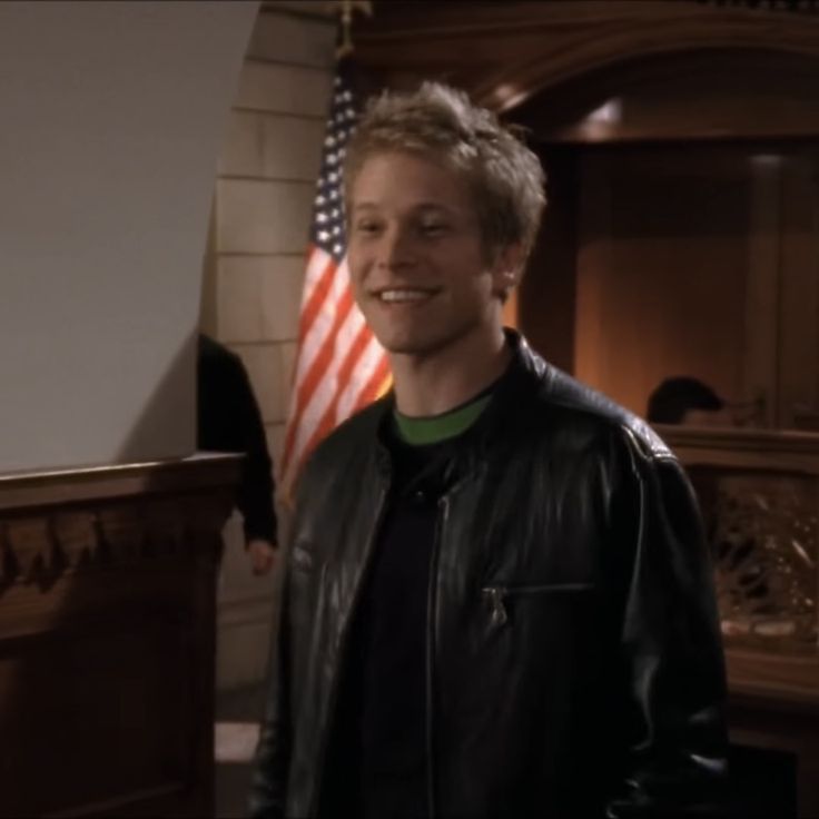 a man standing in front of an american flag wearing a black leather jacket and green t - shirt