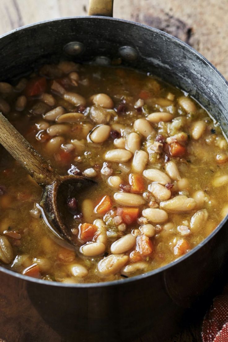 a pot filled with beans and carrots on top of a wooden table next to a spoon