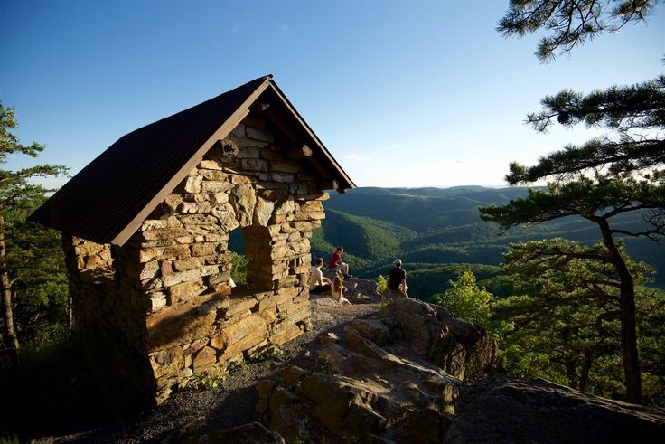 three people standing on top of a rock structure in the woods with mountains in the background